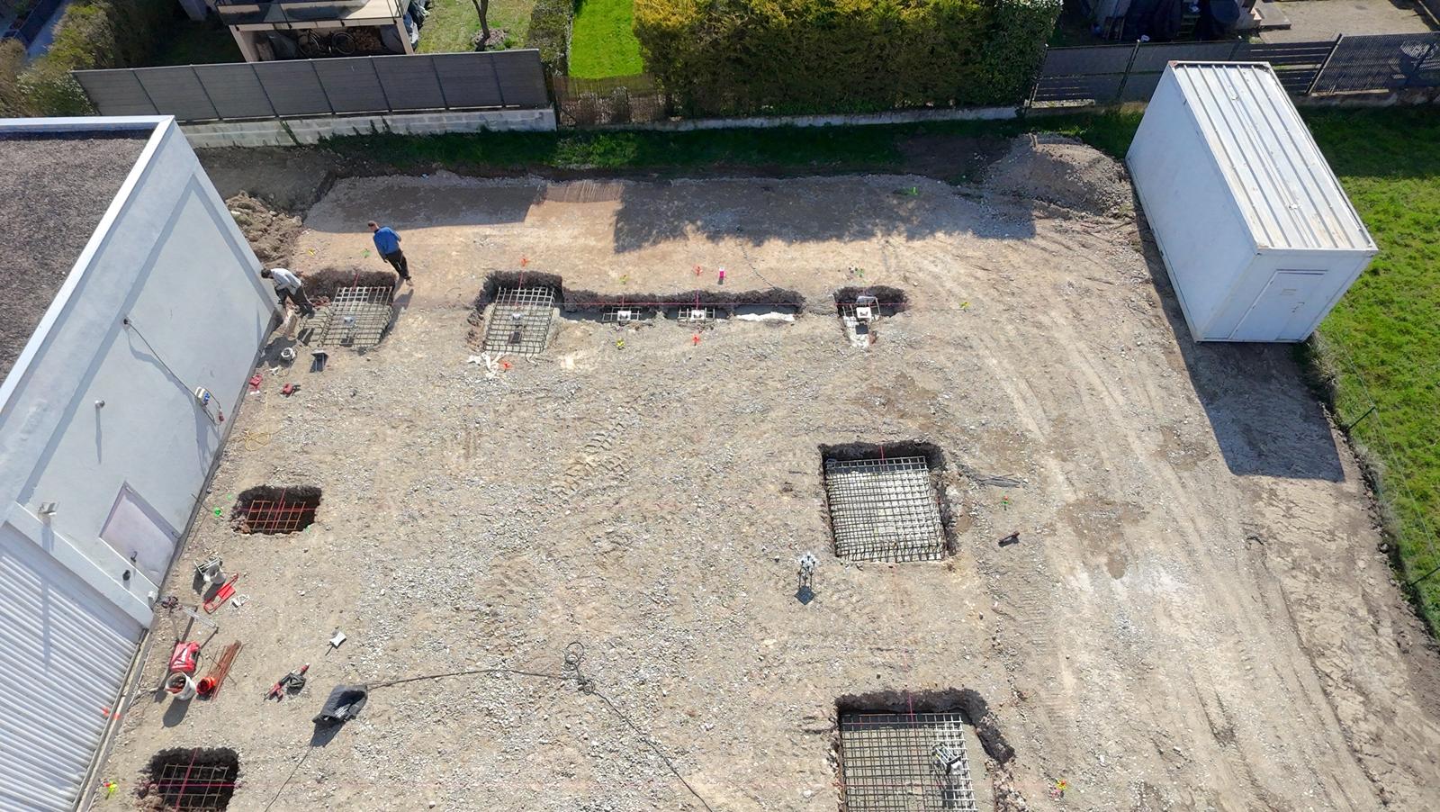 Chantier avec trous creusés et armatures métalliques, deux ouvriers travaillent près d'un mur blanc.