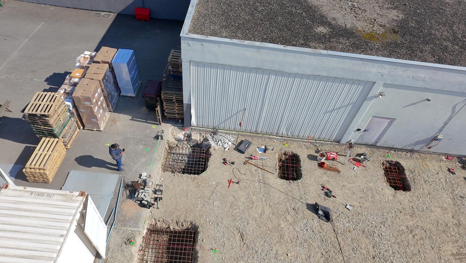 Vue aérienne d'un chantier avec des fondations creusées et une personne debout près de palettes et cartons.