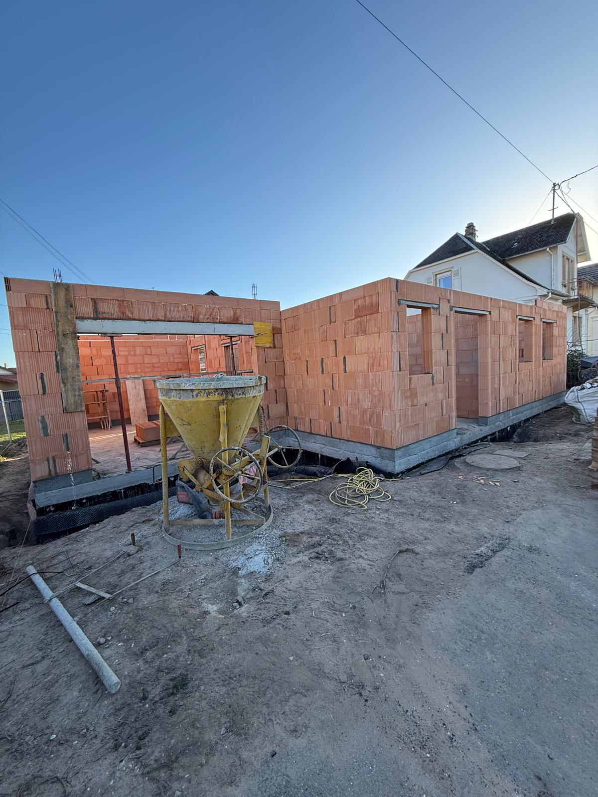 Chantier avec murs non finis en briques rouges et un équipement de construction jaune au premier plan.
