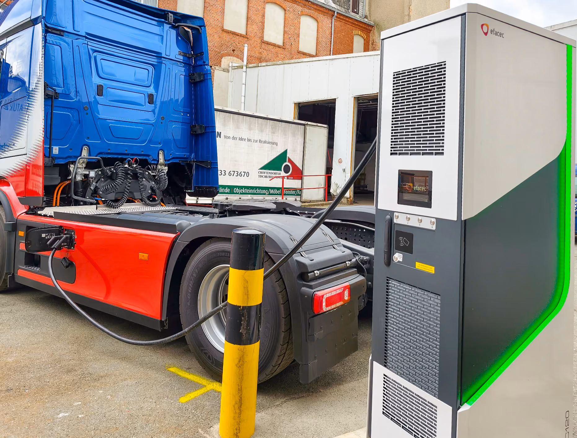 Ein Elektro-Lkw ist an einer Ladestation angeschlossen, umgeladen mit blauem Fahrerhaus und rotem Unterteil, geparkt auf einem Gelände vor einem Gebäude.