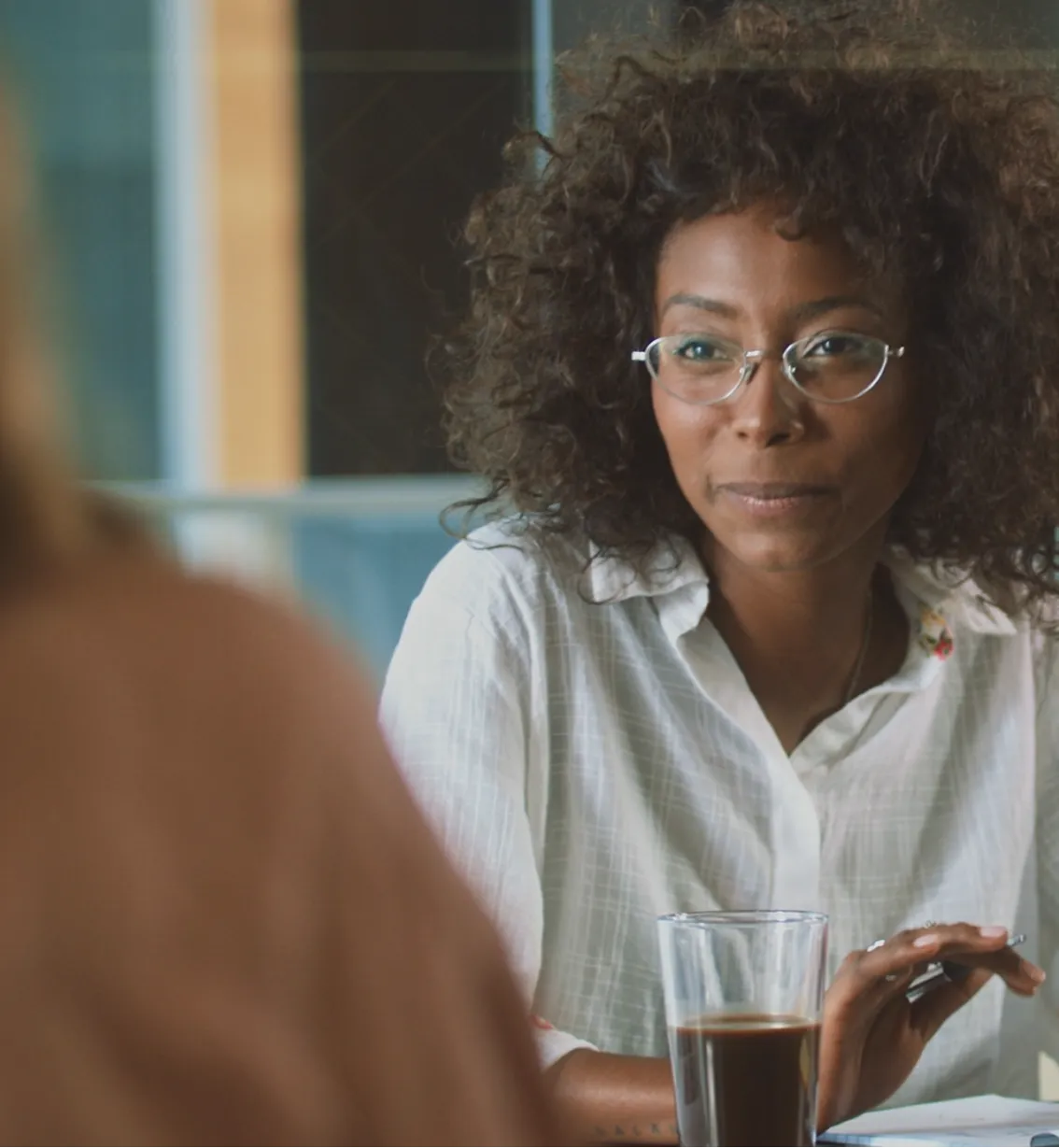 Woman with curly hair and glasses holding a pen with a glass of dark beverage on the table.