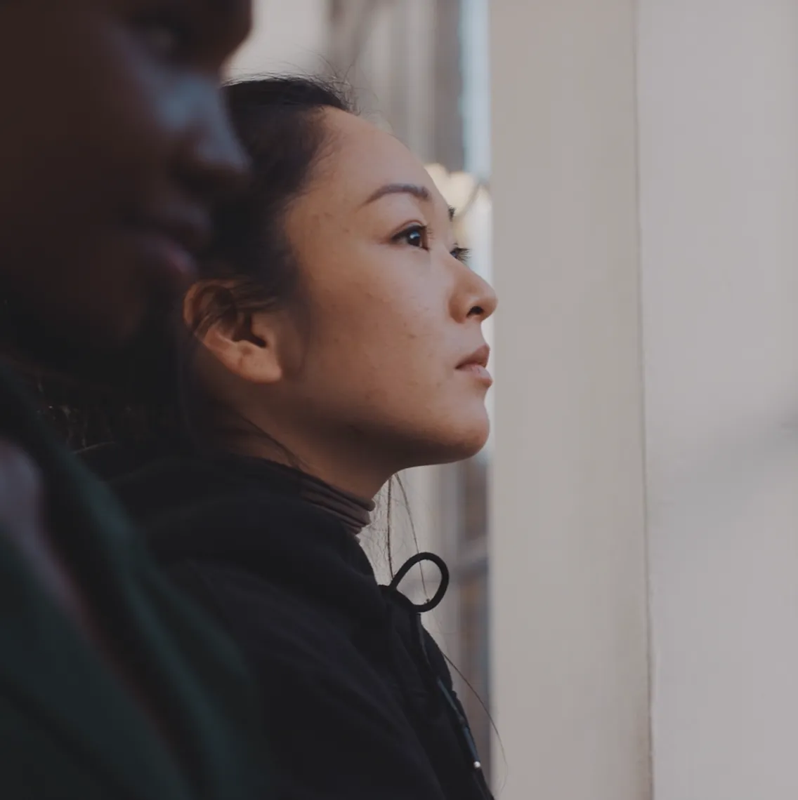 Profile of a woman looking thoughtfully out a window with a blurred figure beside her.