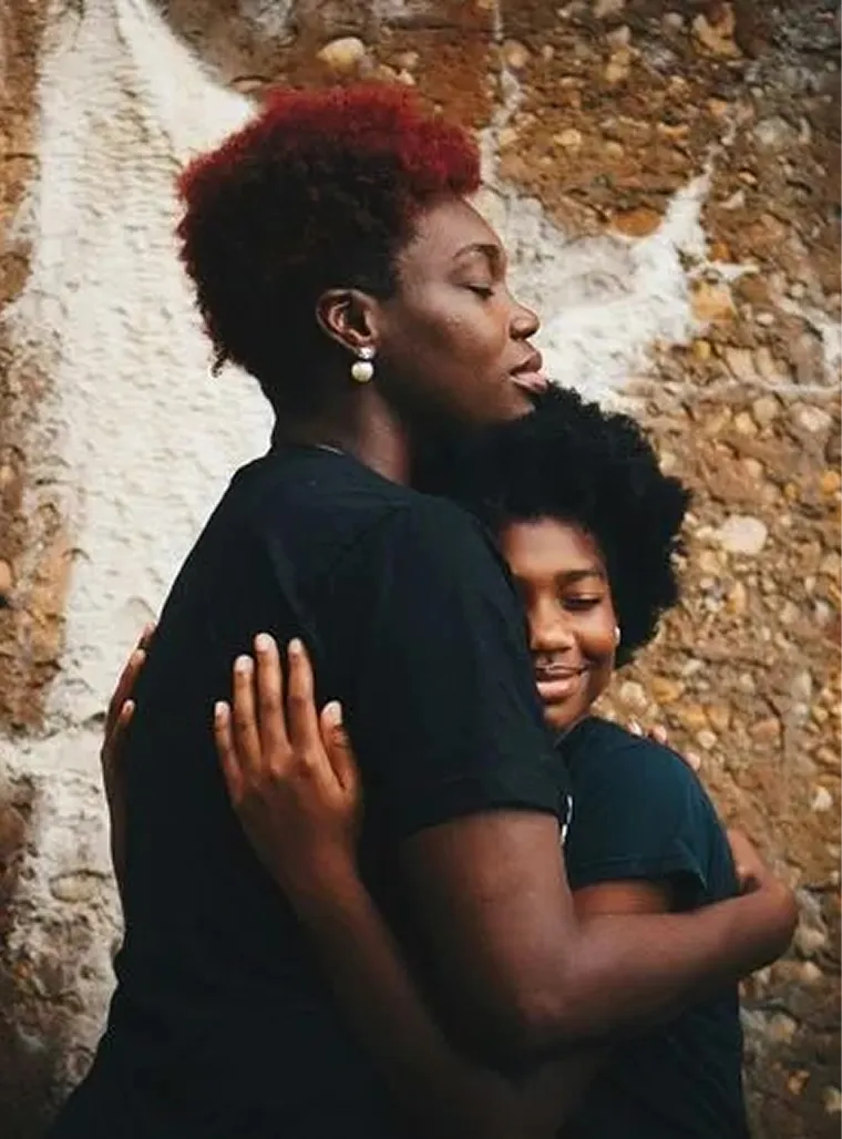 Two people with dark skin and curly hair hugging, one smiling with eyes closed, against a textured wall.