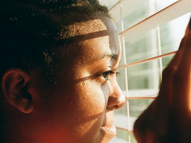 Close-up of a person looking thoughtfully through partially open window blinds with sunlight on their face.