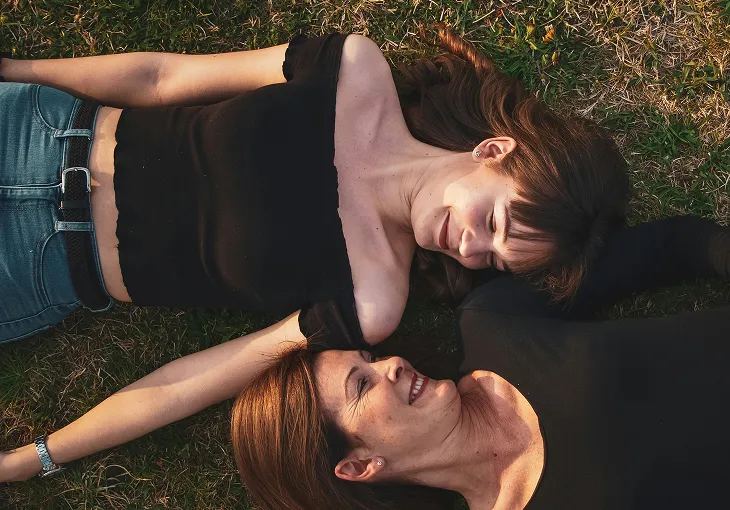 Two women lying on grass, smiling and facing each other, wearing black tops and jeans.