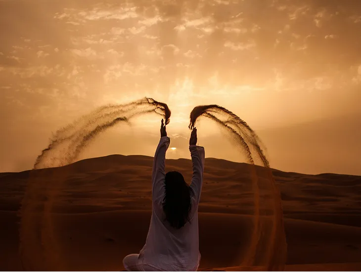 Person in white throws sand into the air at sunset in a desert with sand dunes.