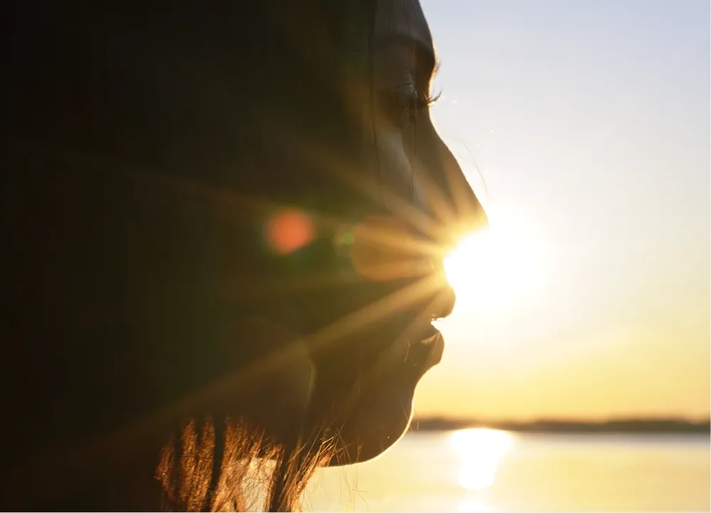 Silhouette of a person in profile with sun rays shining through their face at sunset by water.