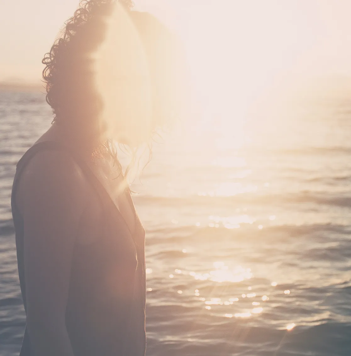 Person with curly hair standing by water as sun shines brightly creating lens flare.
