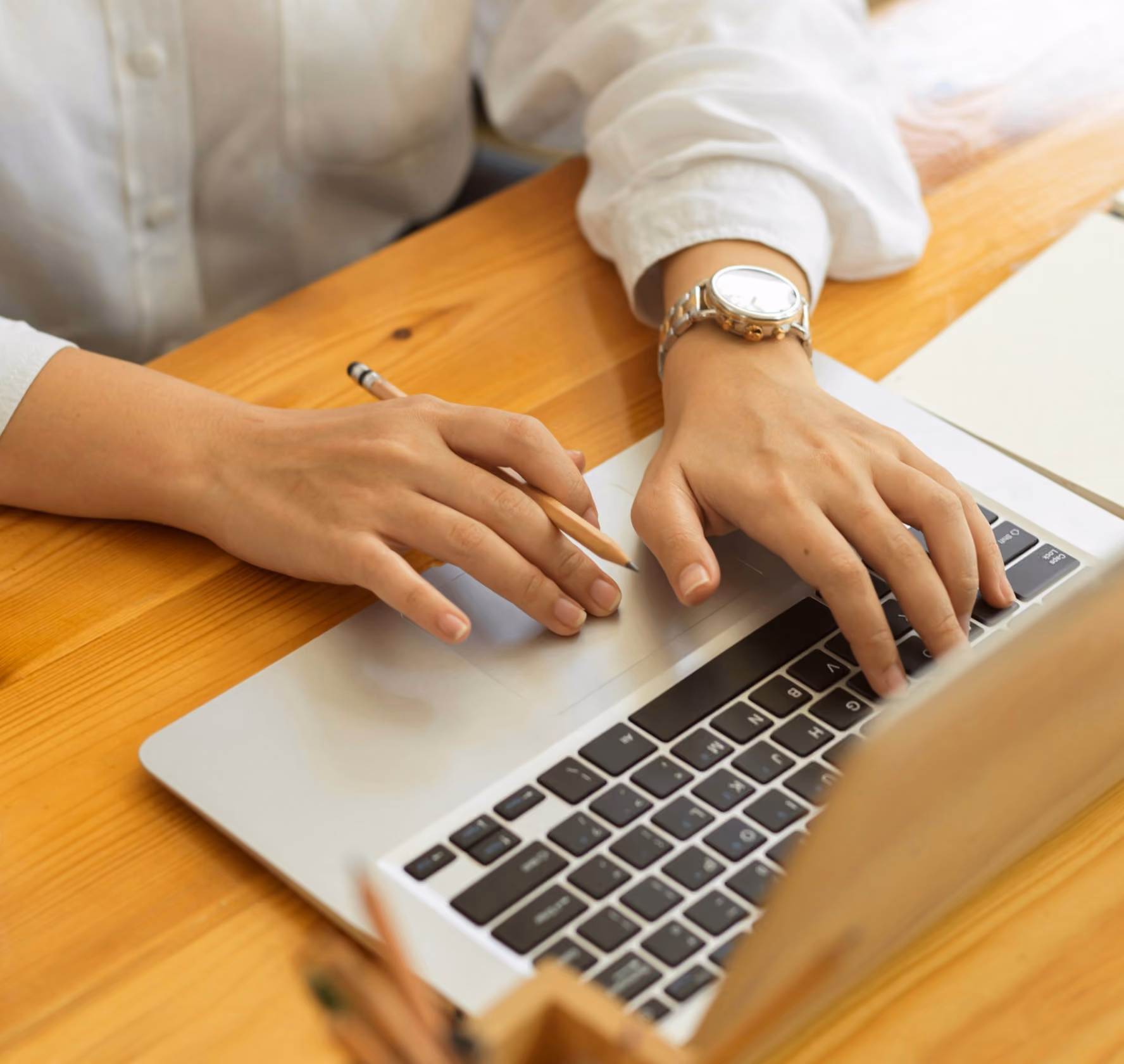 Person typing on a laptop keyboard with one hand while holding a pencil in the other, sitting at a wooden table.