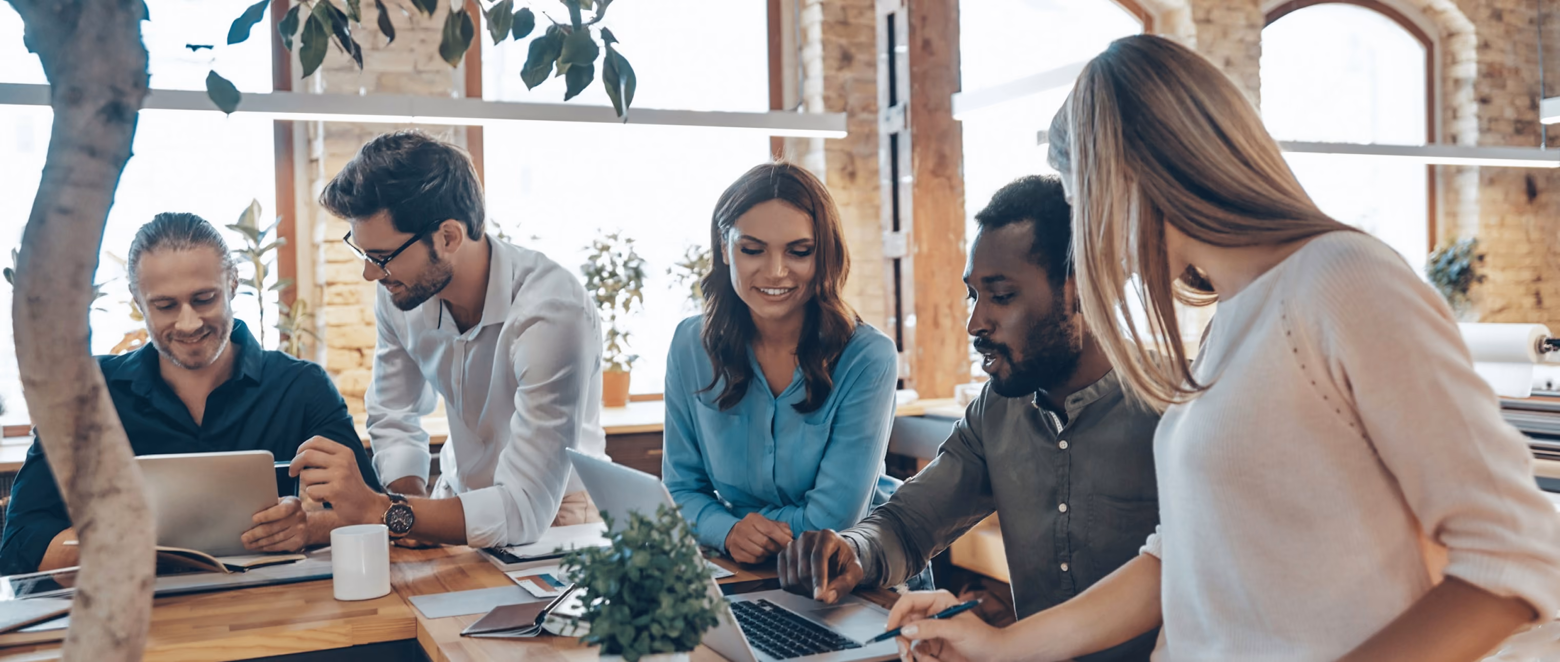Five diverse coworkers collaborating and smiling around a table with laptops and documents in a bright modern office with large windows and plants.