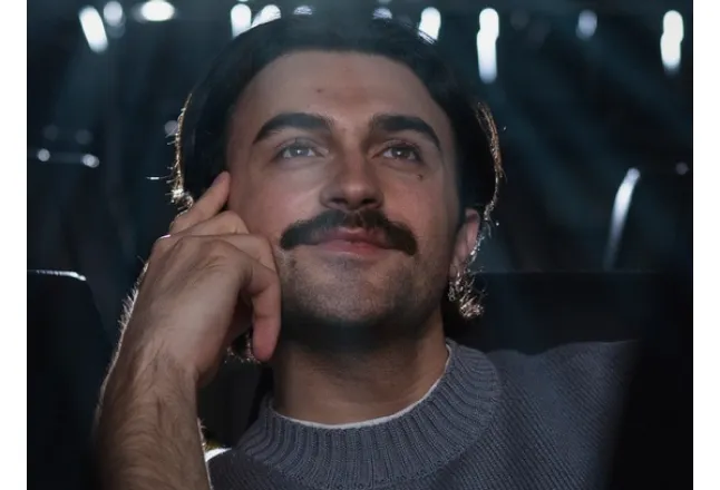 Man with mustache and sweater seated in dim light looking thoughtfully upward.