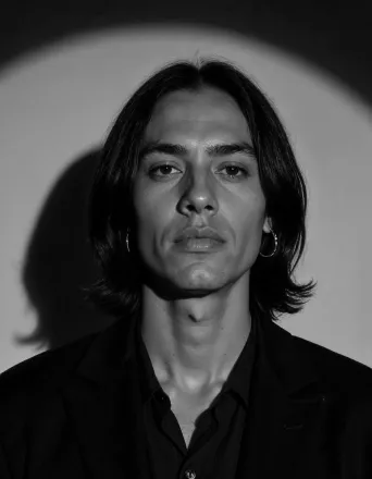 Black-and-white portrait of a young person with shoulder-length hair, wearing hoop earrings and a dark shirt.