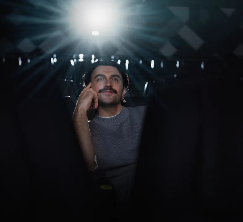 Man with mustache sitting alone in dark theater, light shining behind him.