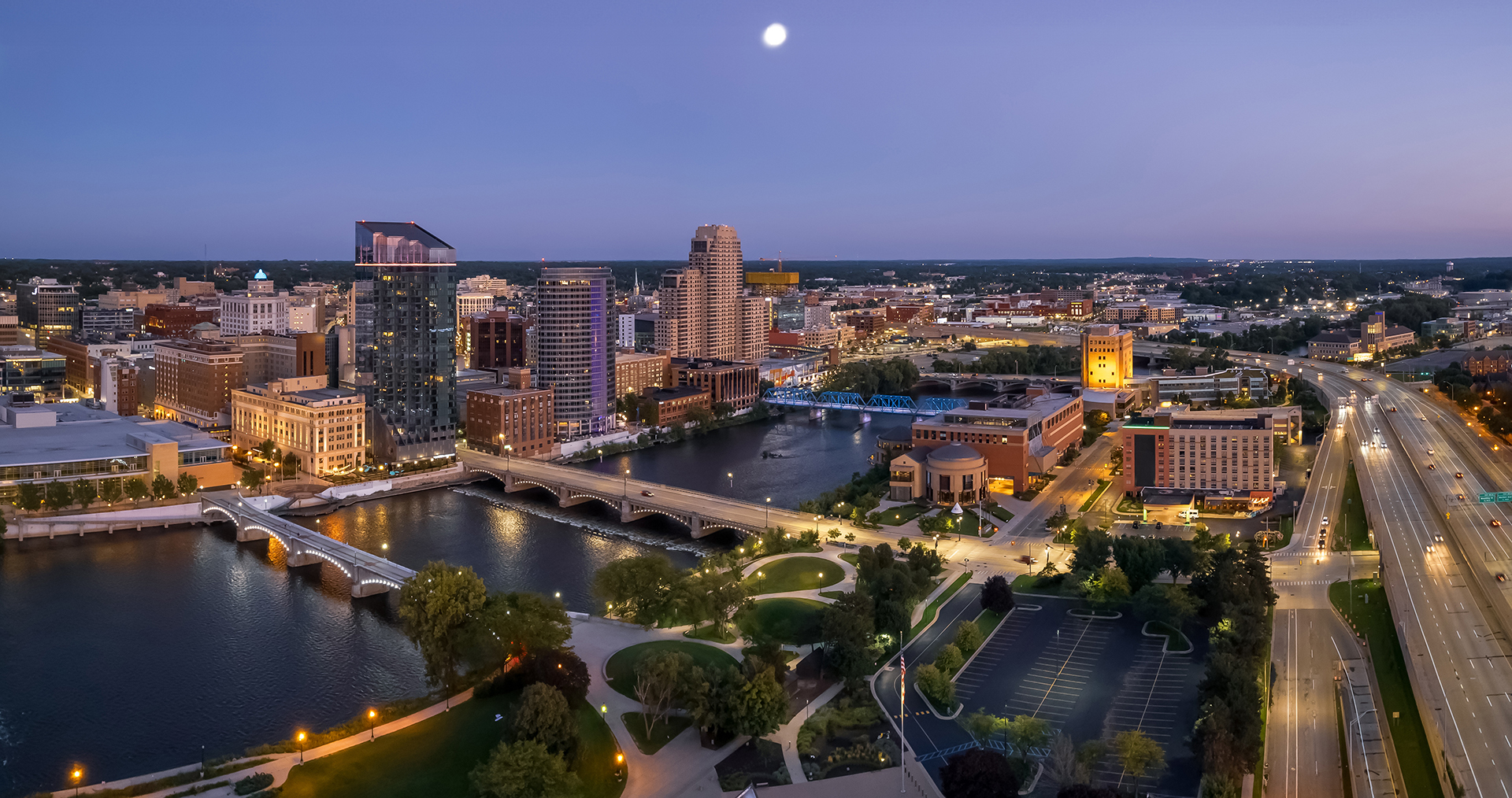 Aerial view of a Grand Rapids, MI at dusk with illuminated bridges over a river and lit buildings.