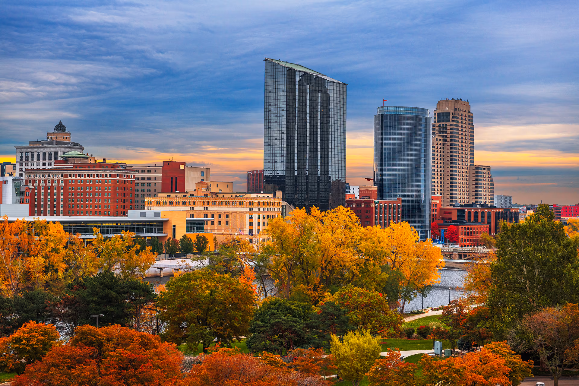 Grand Rapids city skyline with glass skyscrapers and colorful autumn trees by a river under a cloudy sky.