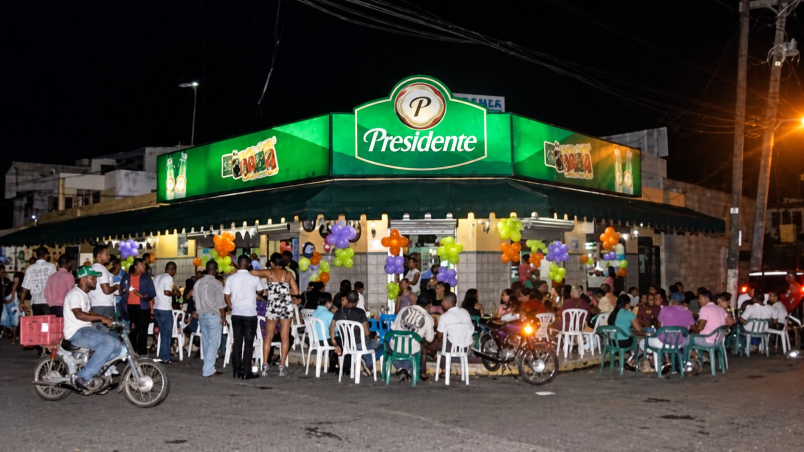 Personas reunidas de noche frente a un colmadón, con mesas, sillas y motocicletas en la calle.