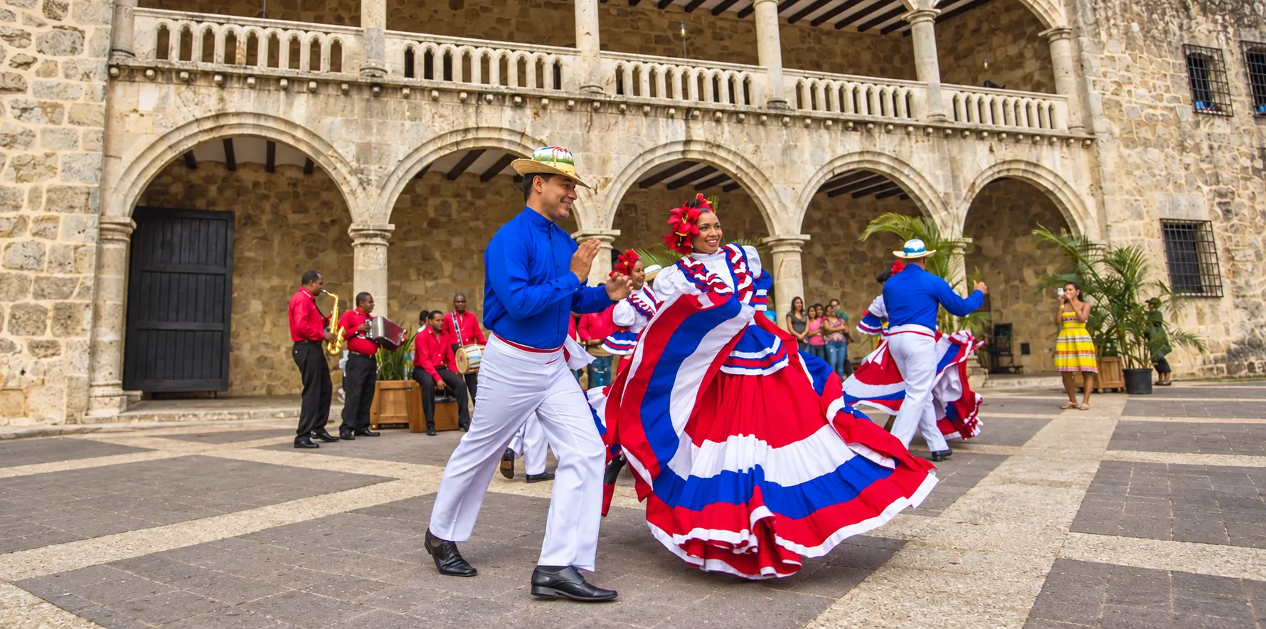 Pareja con vestuario rojo, blanco y azul bailando frente a una fachada de piedra con arcos.