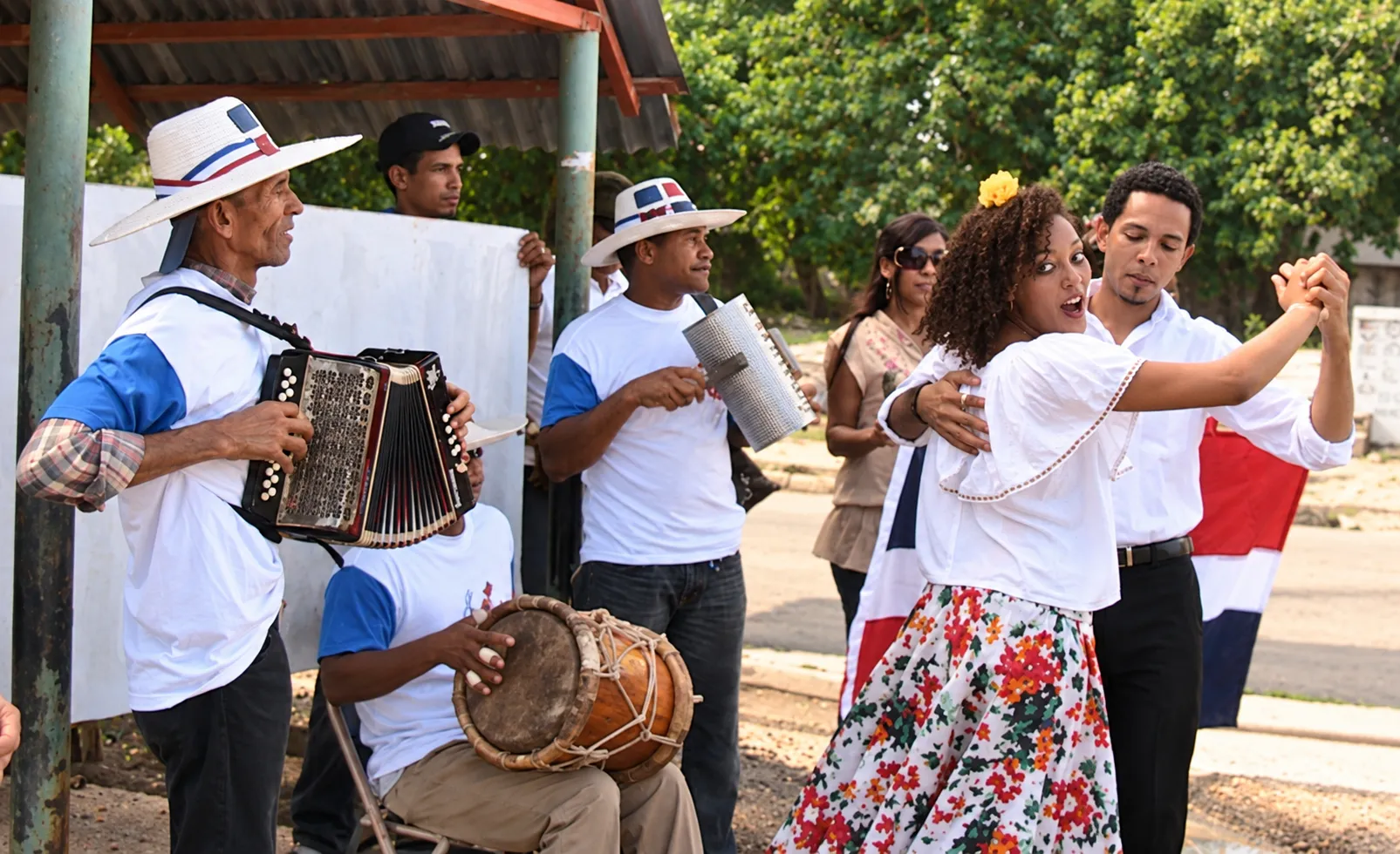 Pareja bailando al aire libre junto a músicos con acordeón, güira y tambora.