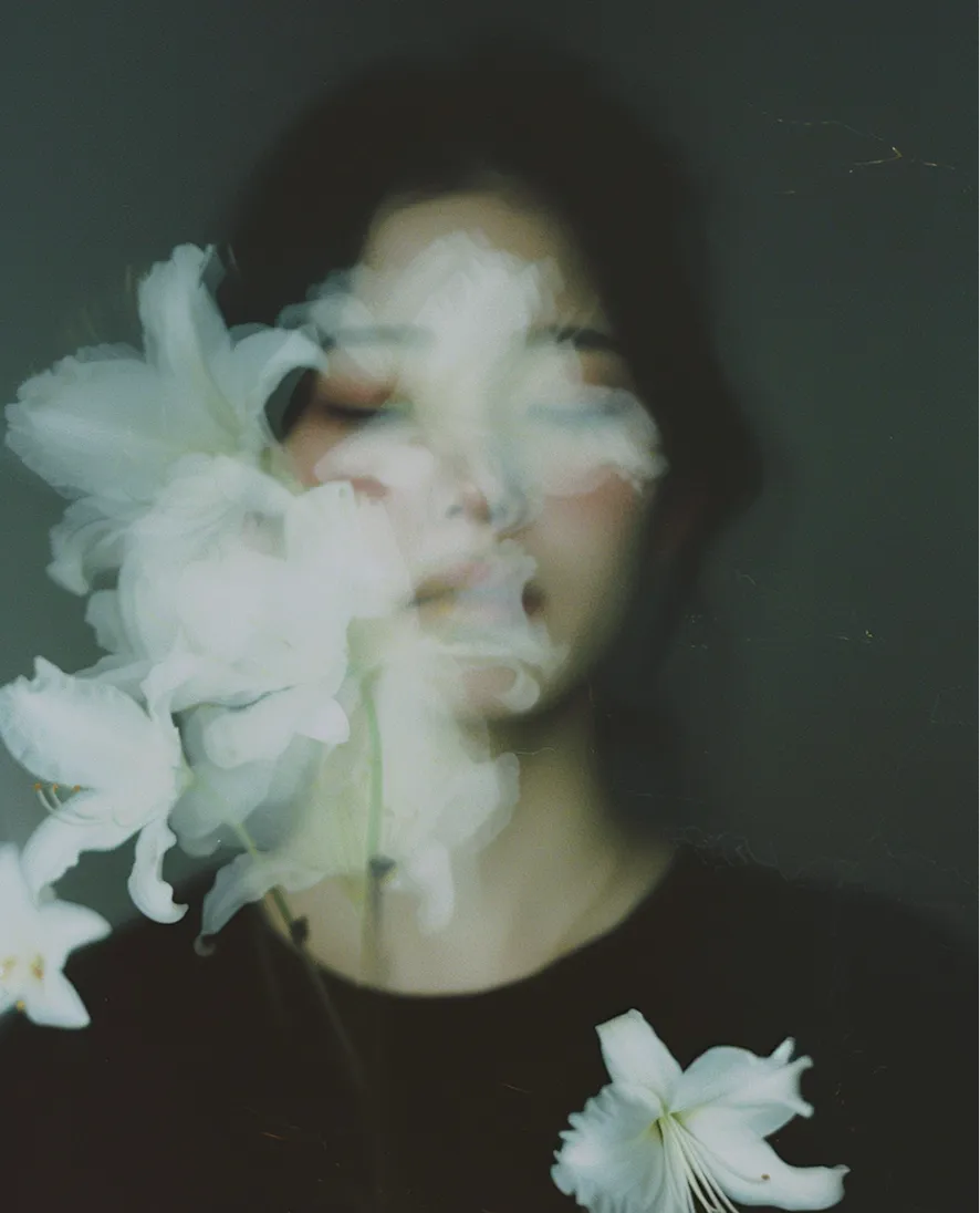 Blurred portrait of a person with closed eyes holding white lilies against a dark background.