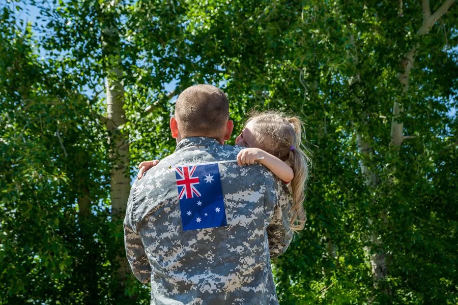 Soldier in camouflage uniform with Australian flag on back holding a young girl with ponytail outdoors.