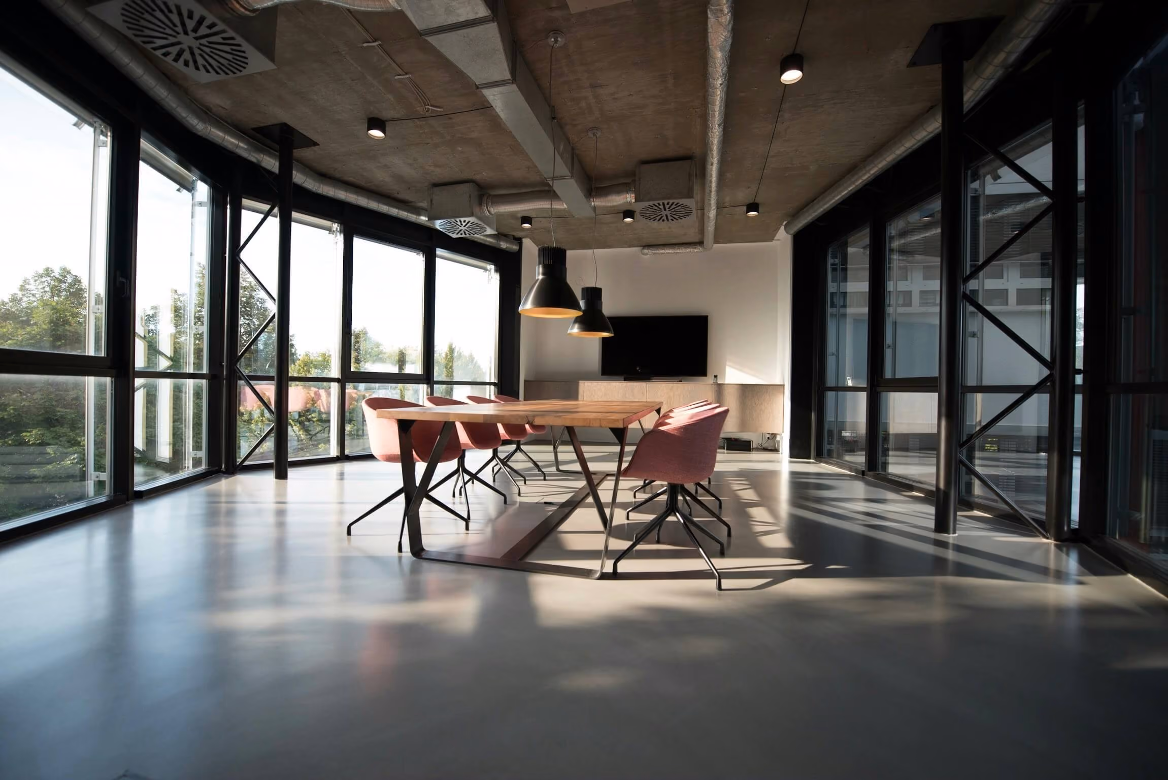 Modern conference room with large windows, wooden table, pink chairs, and two black pendant lights.