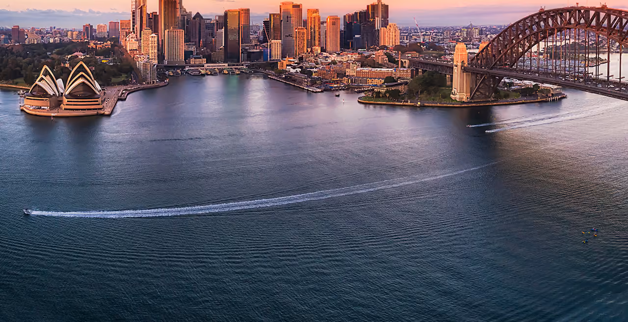 Aerial view of Sydney Opera House, Harbor Bridge, and city skyline at sunset with boats in water.
