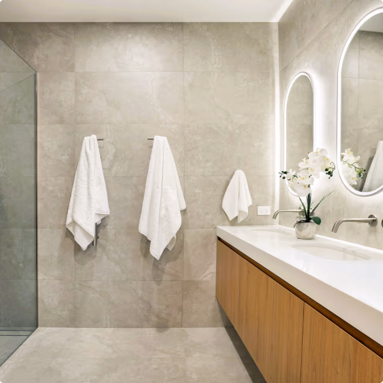 Modern bathroom with gray tiled walls, three white towels, a double sink, oval mirrors, and a flower vase.