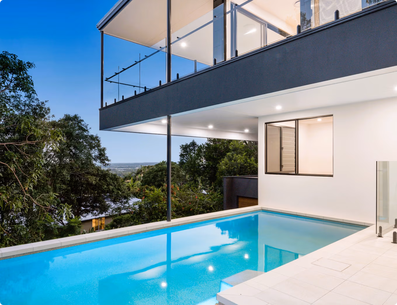 Modern house with a glass balcony overlooks a blue outdoor swimming pool surrounded by trees.