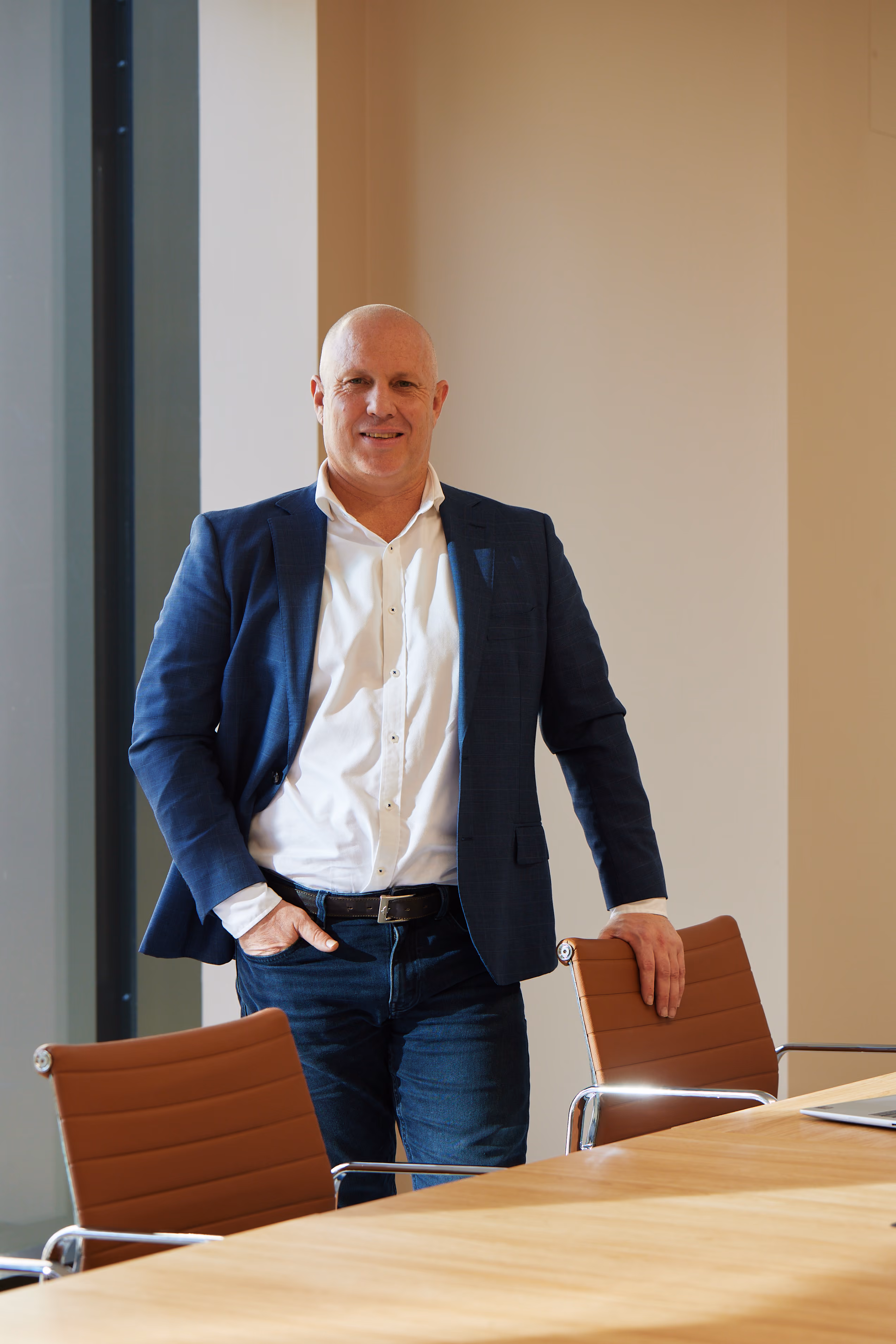 Bald man in a navy blazer and white shirt stands smiling in a modern office by a wooden table.