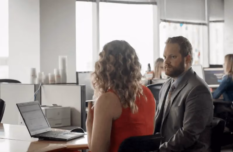 Man in a suit and woman with curly hair in a red dress talking in a modern office with laptops.