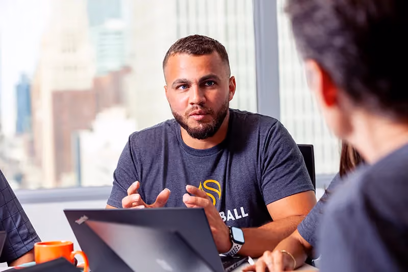 Man with beard talking and gesturing at a meeting with laptop and city buildings in background.