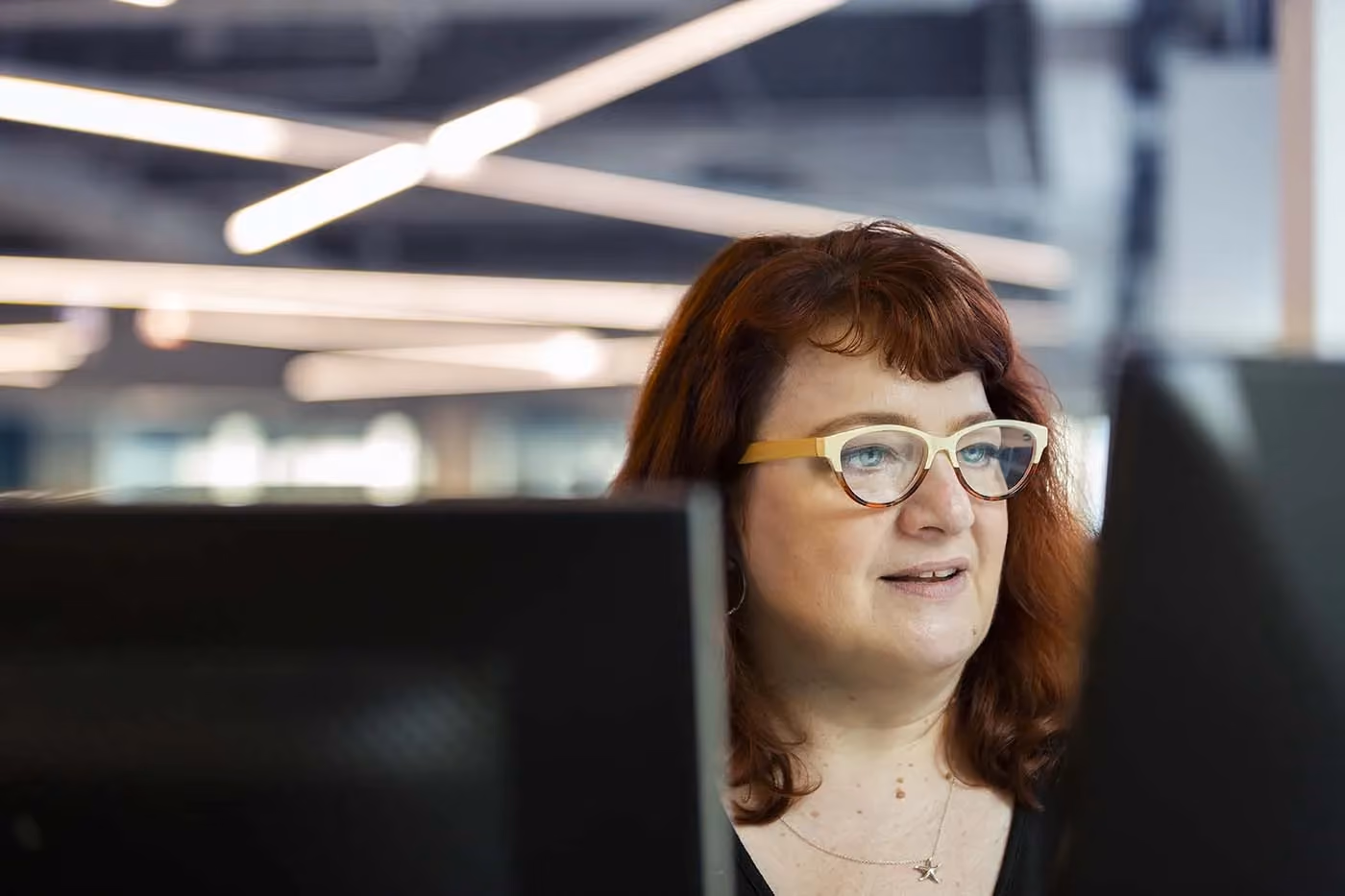 Woman with red hair and glasses looking at two computer monitors in an office setting.