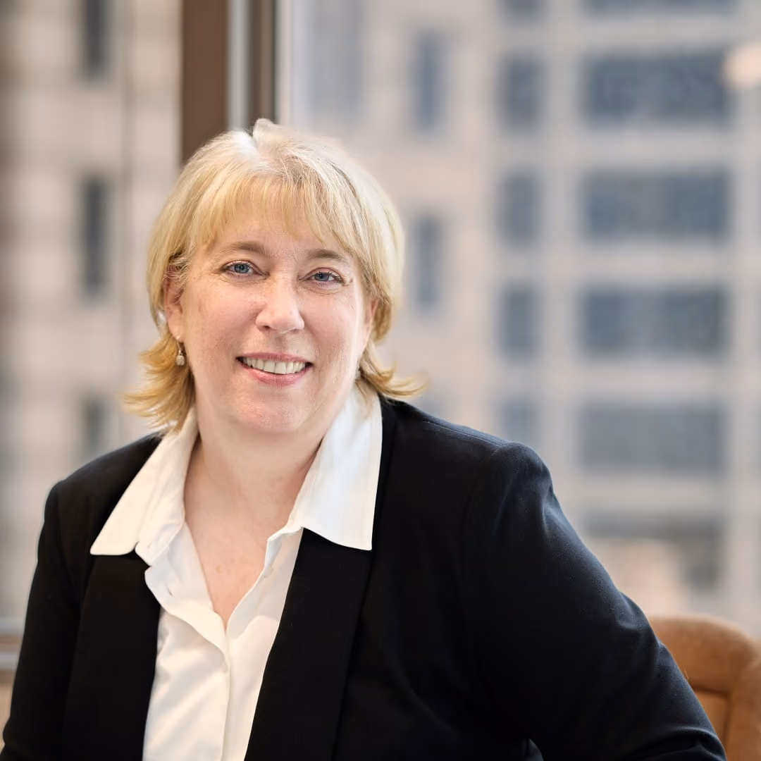 Smiling woman with short blonde hair wearing a white shirt and black blazer in an office setting.