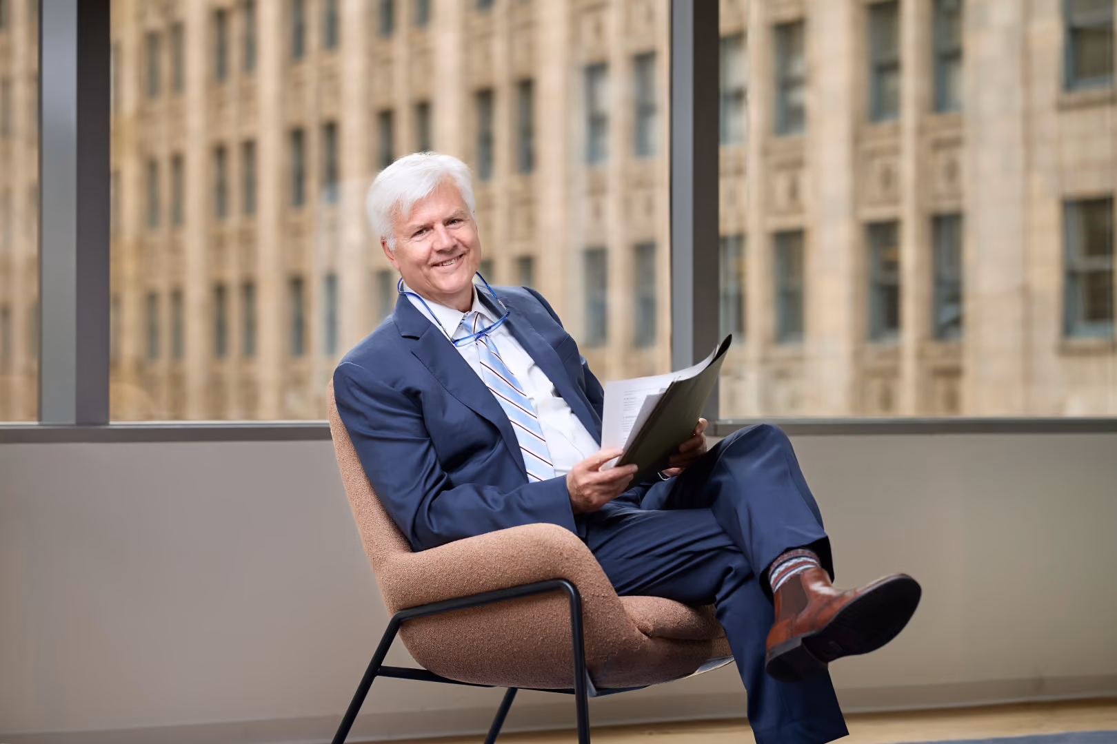 Smiling white-haired man in a blue suit sitting cross-legged in a chair holding papers.