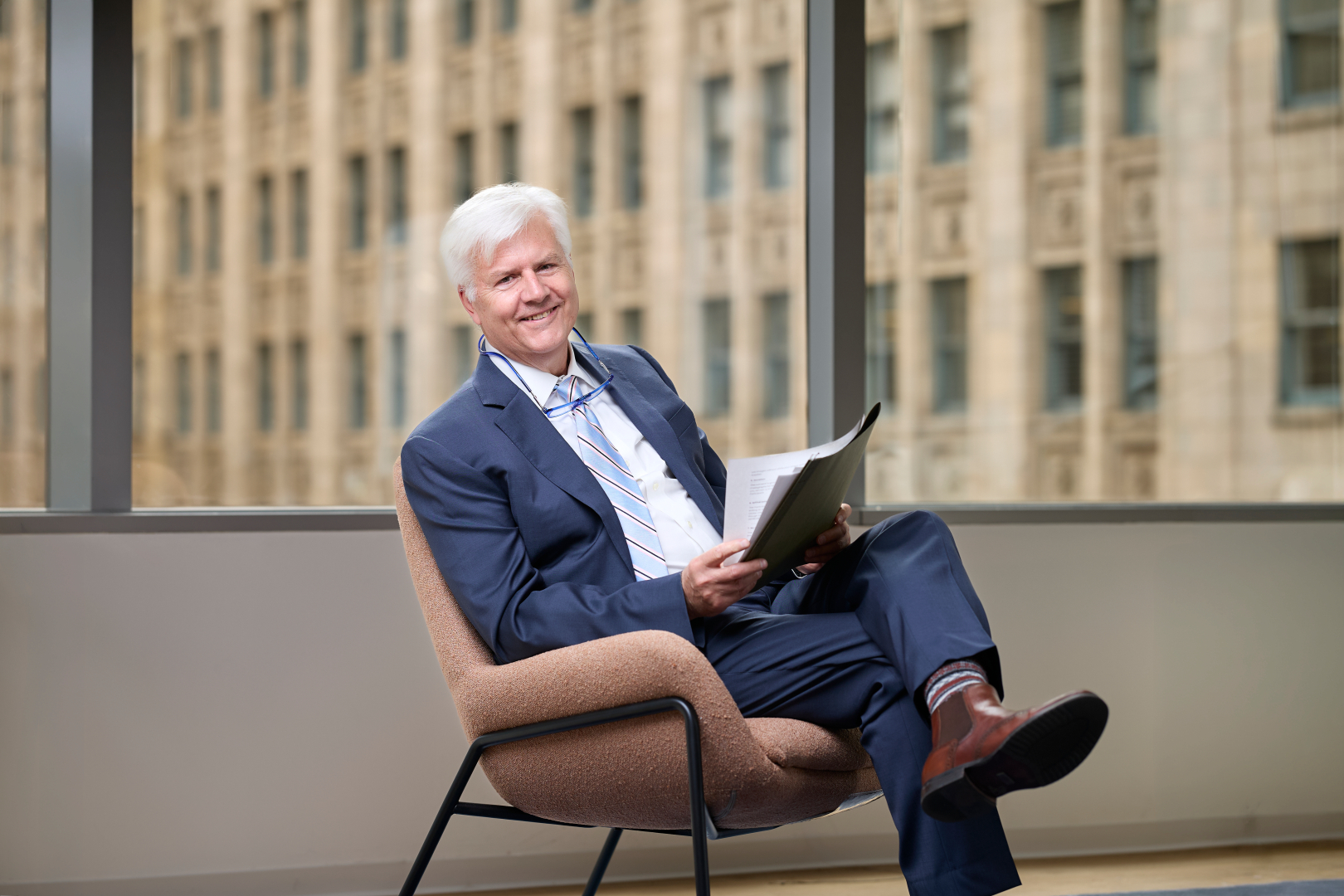 Smiling white-haired man in a blue suit sitting cross-legged in a chair holding papers.