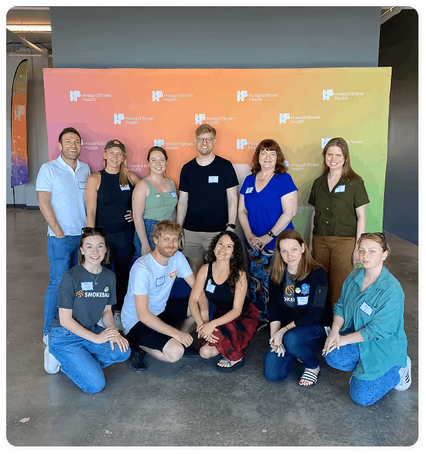 Group of eleven adults posing indoors in front of a Howard Brown Health rainbow backdrop.