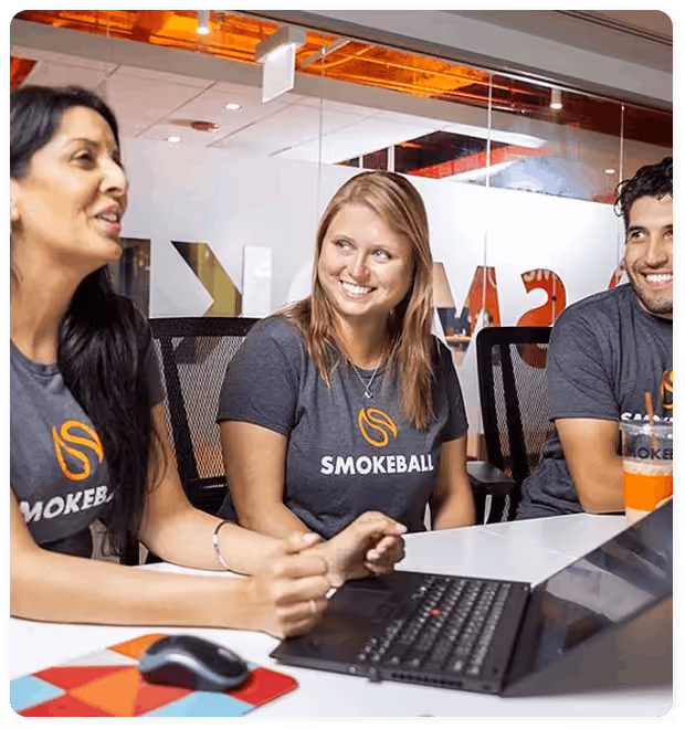 Three people smiling and talking at a desk, wearing matching Smokeball t-shirts with a laptop and mouse on the table.