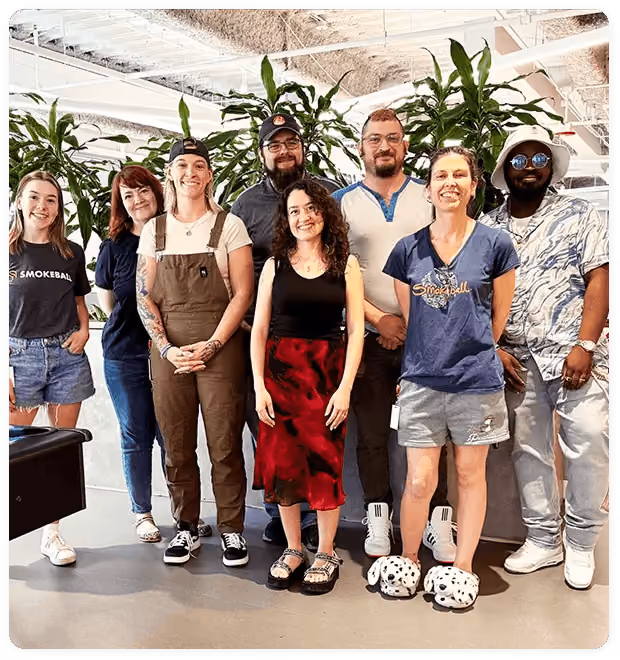 Eight diverse people standing together indoors with plants in the background, smiling at the camera.
