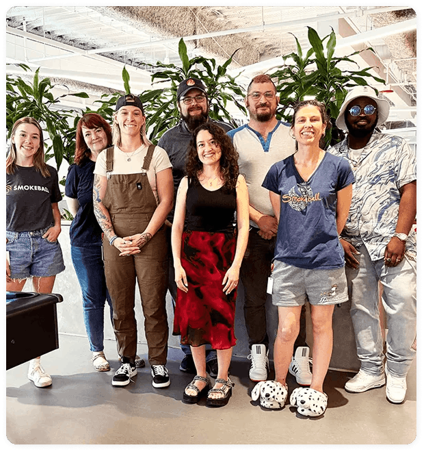 Eight diverse people standing together indoors with plants in the background, smiling at the camera.