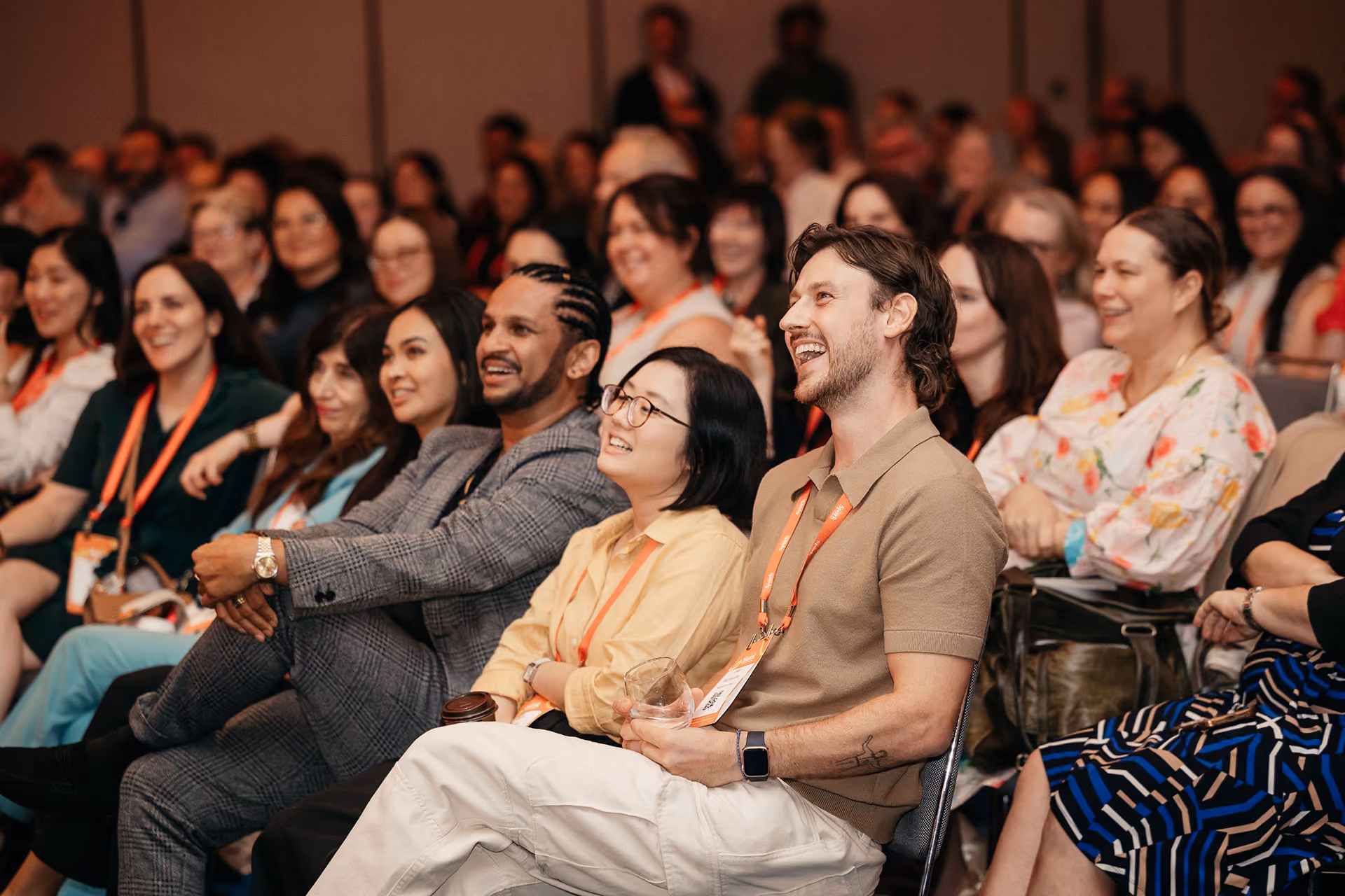 Audience of diverse people smiling and laughing while seated at an indoor event or conference.