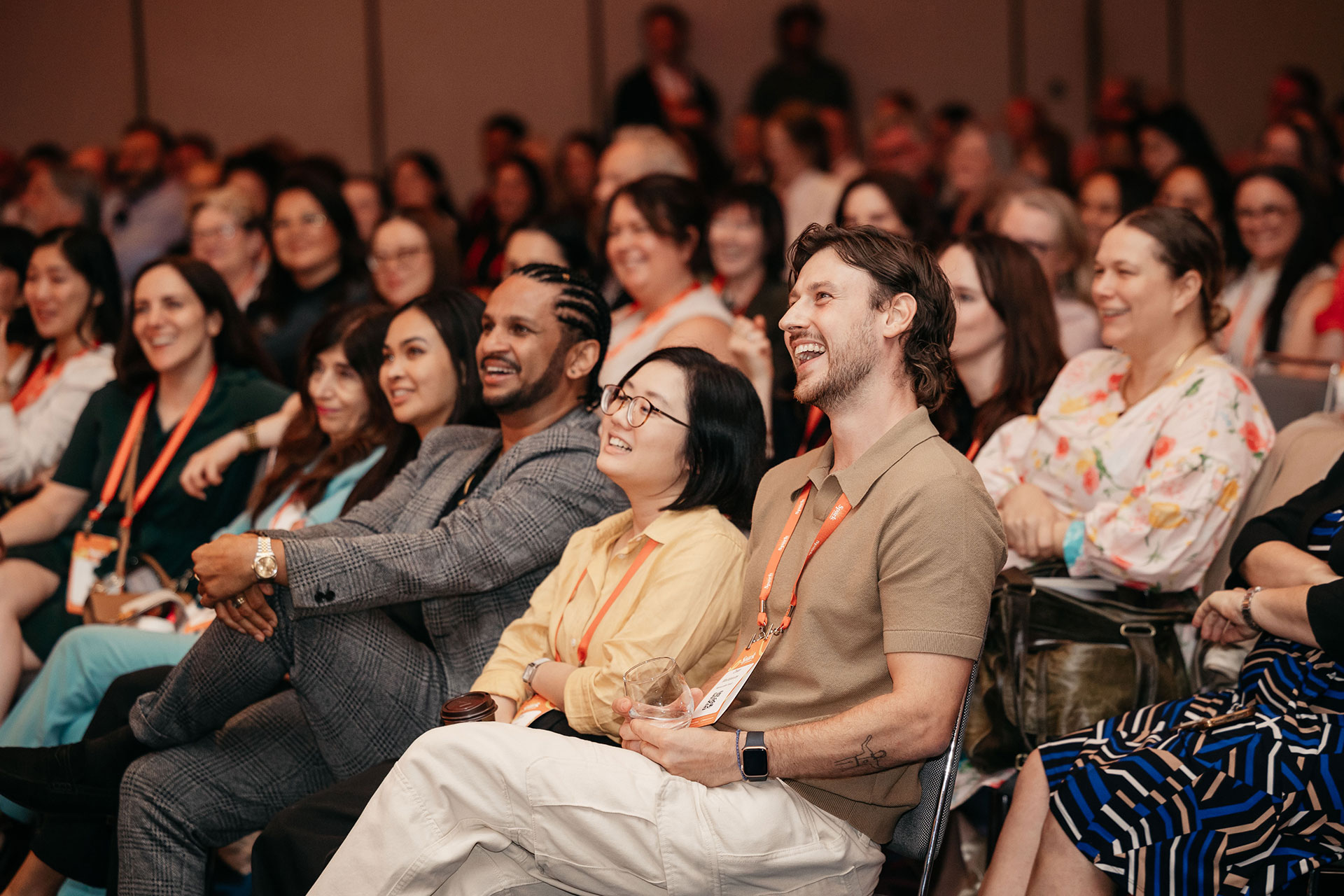 Audience of diverse people smiling and laughing while seated at an indoor event or conference.
