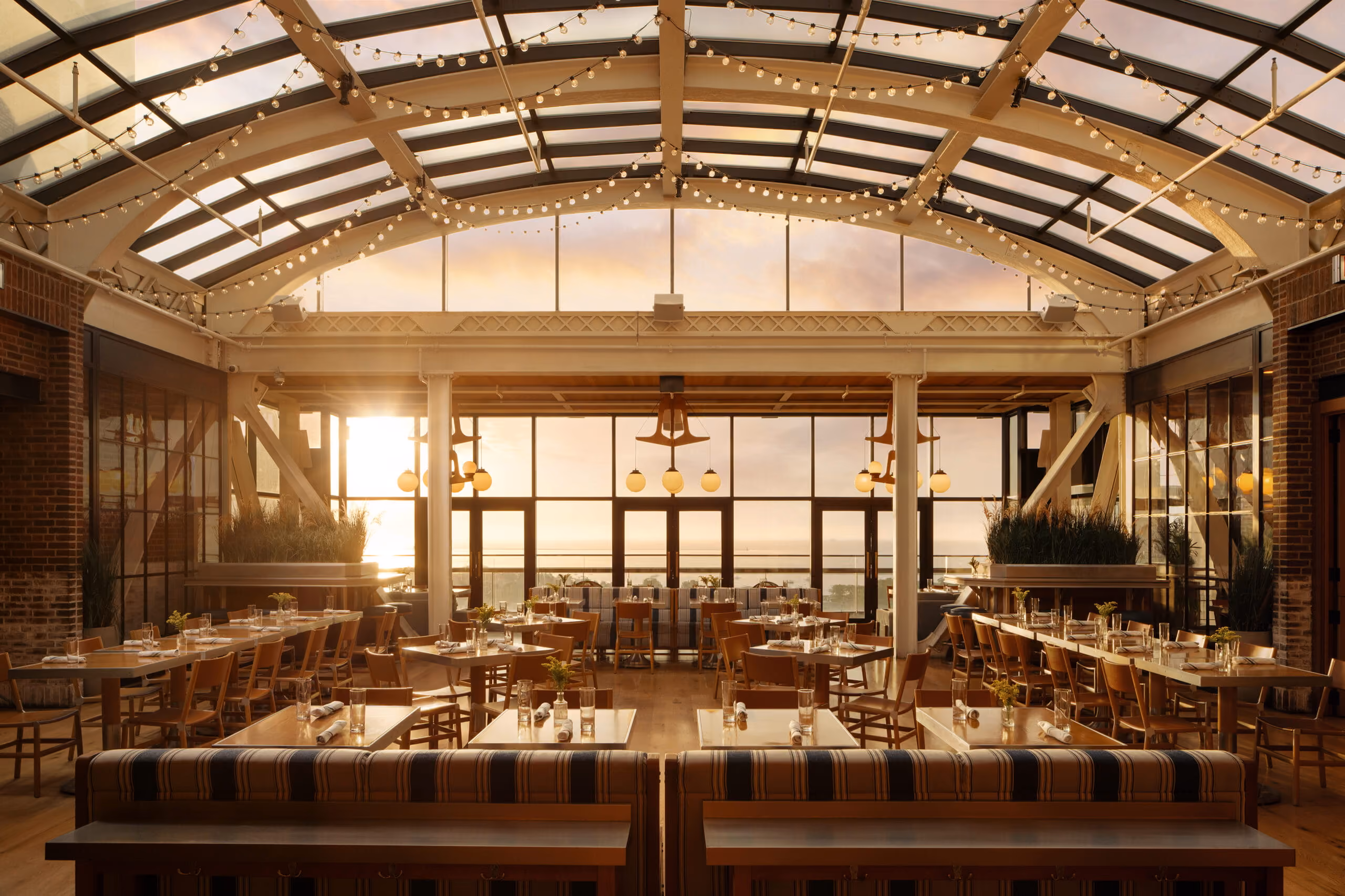 Sunlit rooftop restaurant with wooden tables, chairs, string lights, and large windows overlooking water.