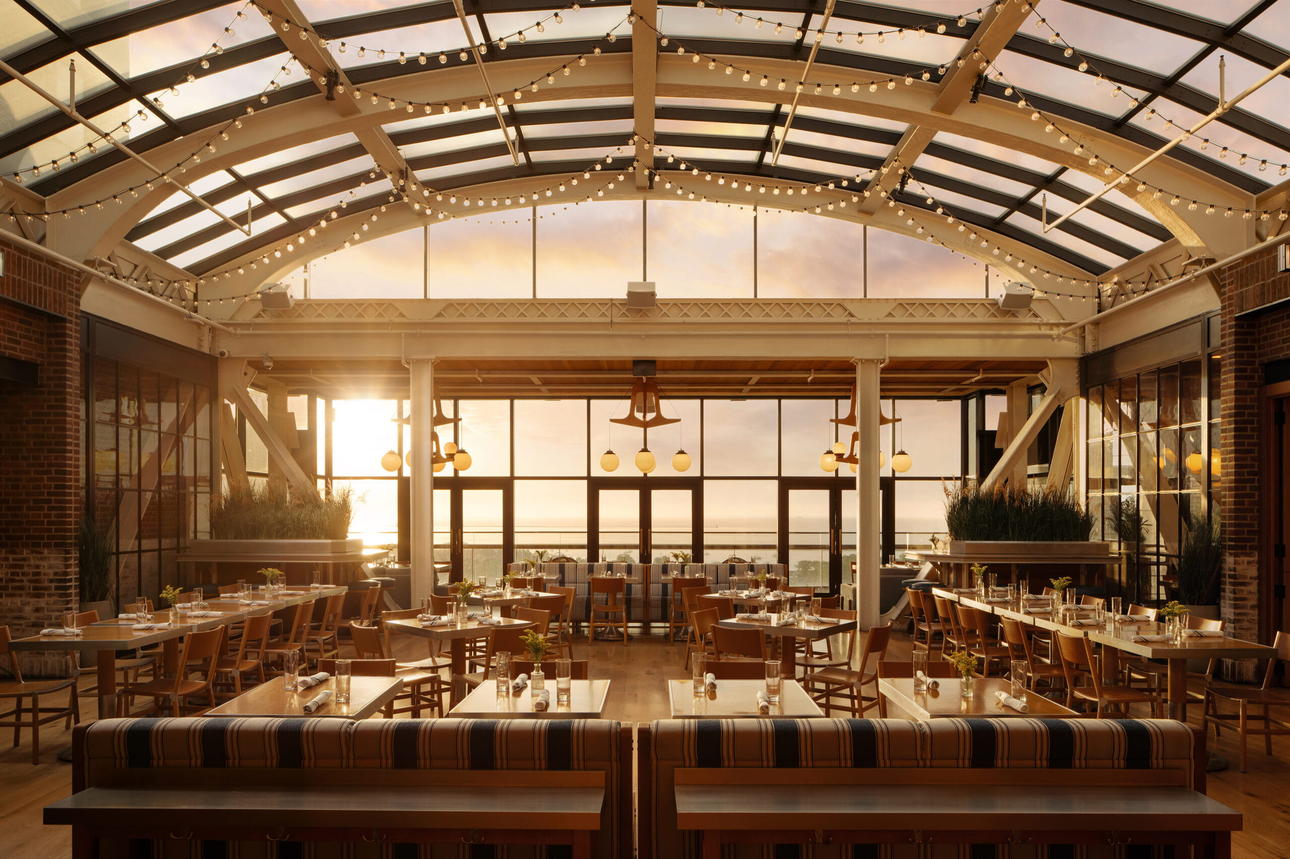 Sunlit rooftop restaurant with wooden tables, chairs, string lights, and large windows overlooking water.