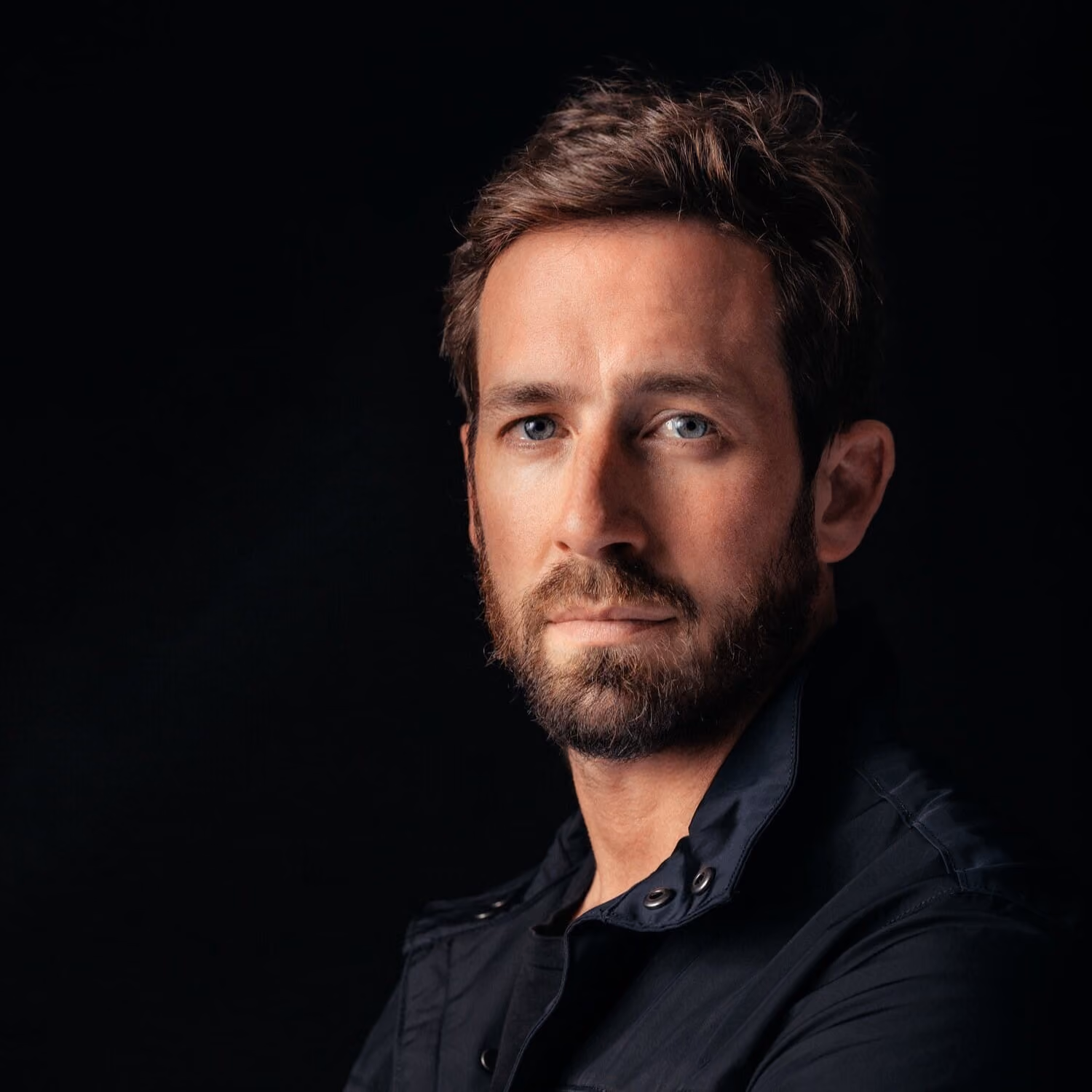 Serious bearded man with blue eyes wearing a dark jacket against a black background.