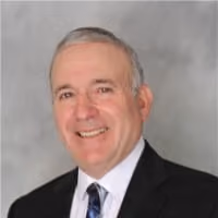 Smiling middle-aged man in business suit with white shirt and patterned tie against gray background.