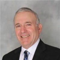 Smiling middle-aged man in business suit with white shirt and patterned tie against gray background.