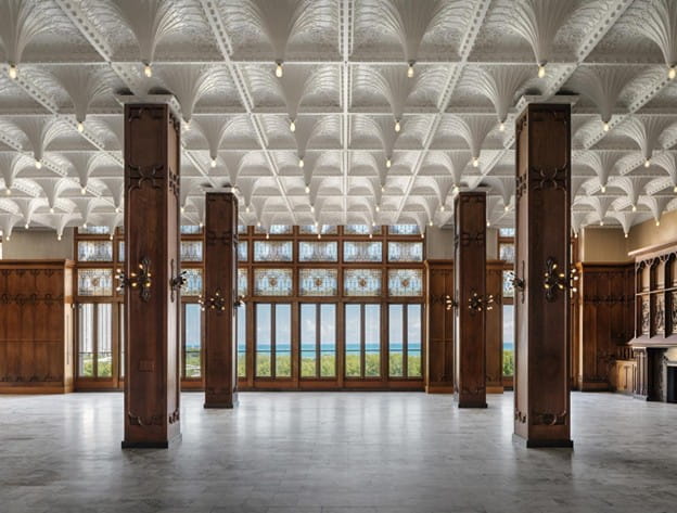 Spacious hall with decorative wood columns, intricate white ceiling, and large windows overlooking green landscape.