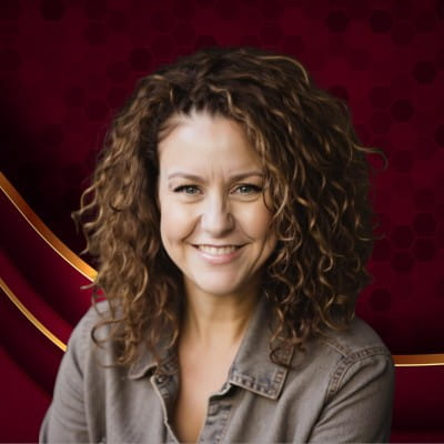 Smiling woman with curly brown hair wearing a gray buttoned shirt against a dark red background.