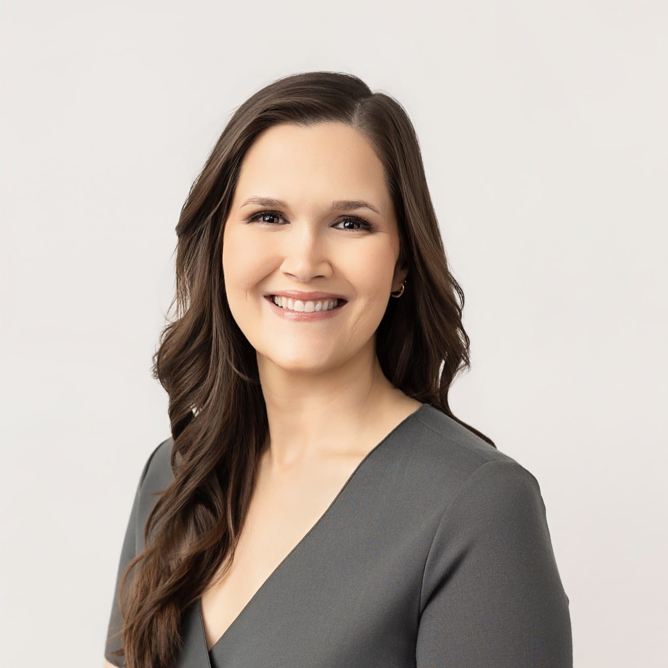Smiling woman with long brown hair wearing a gray top against a plain light background.