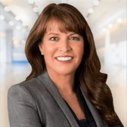 Smiling woman with long brown hair wearing a gray blazer in a bright office space.